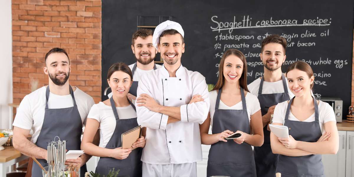 A smiling group of diverse culinary students and kitchen staff standing with their head chef in a training kitchen, showcasing teamwork in a UK culinary arts apprenticeship program.