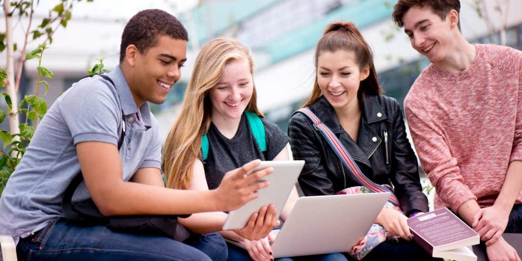 A diverse group of four university students smiling and studying together outdoors on campus with a laptop and digital tablet, representing UK university funding and sanctuary opportunities.