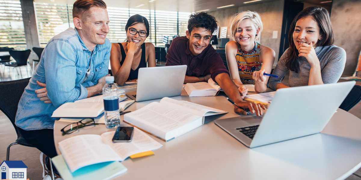 A group of five diverse university students sitting together at a library table, smiling and looking at laptop screens with textbooks open, researching tuition fees and maintenance loans to understand what student finance actually covers.