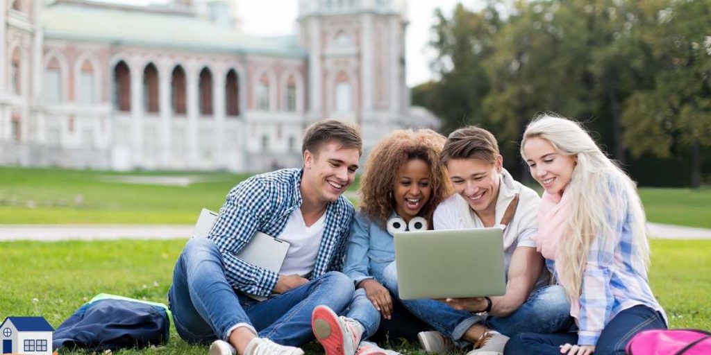 A diverse group of four happy students sitting on the grass outside a university campus, smiling and looking at a laptop to research government student loans, scholarships, and how to apply for university with no money.