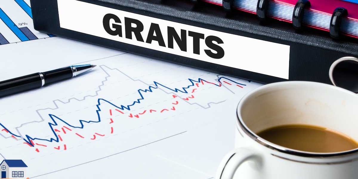 A black office binder with a prominent label reading "GRANTS" sitting on a desk alongside financial charts, a pen, and a cup of coffee, symbolizing budgeting and securing extra university funding.