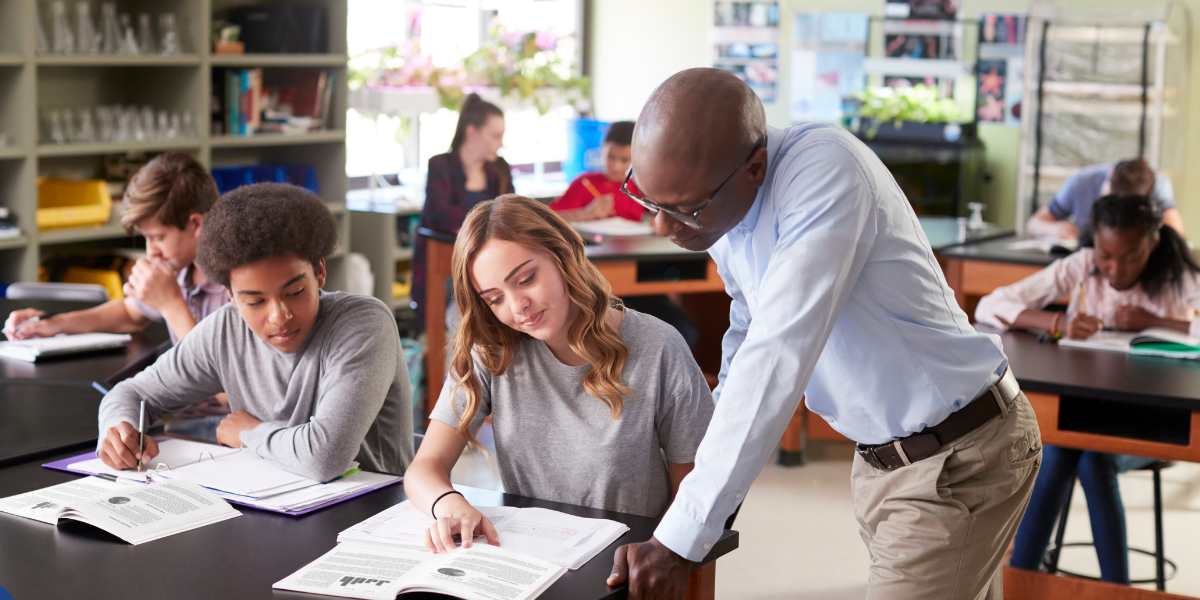 A supportive male teacher leaning over a classroom desk to guide a female high school student through a textbook, with other students studying in a bright classroom setting.