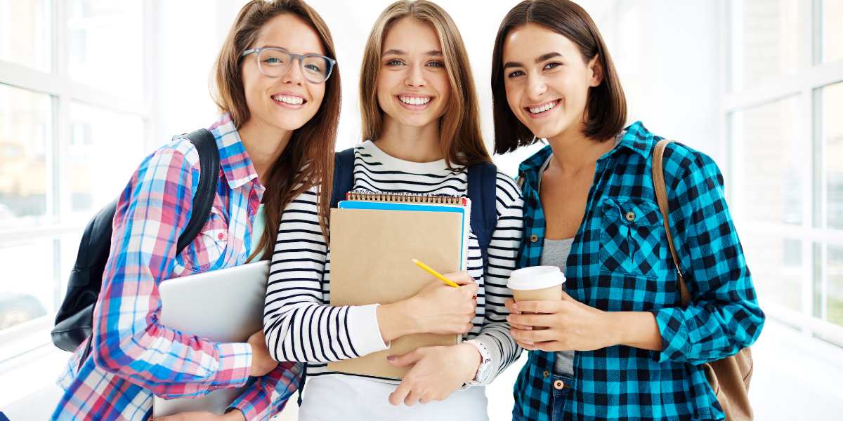 Three cheerful multiethnic female university students standing together in a bright hallway, smiling while holding books, a laptop, and a coffee cup.