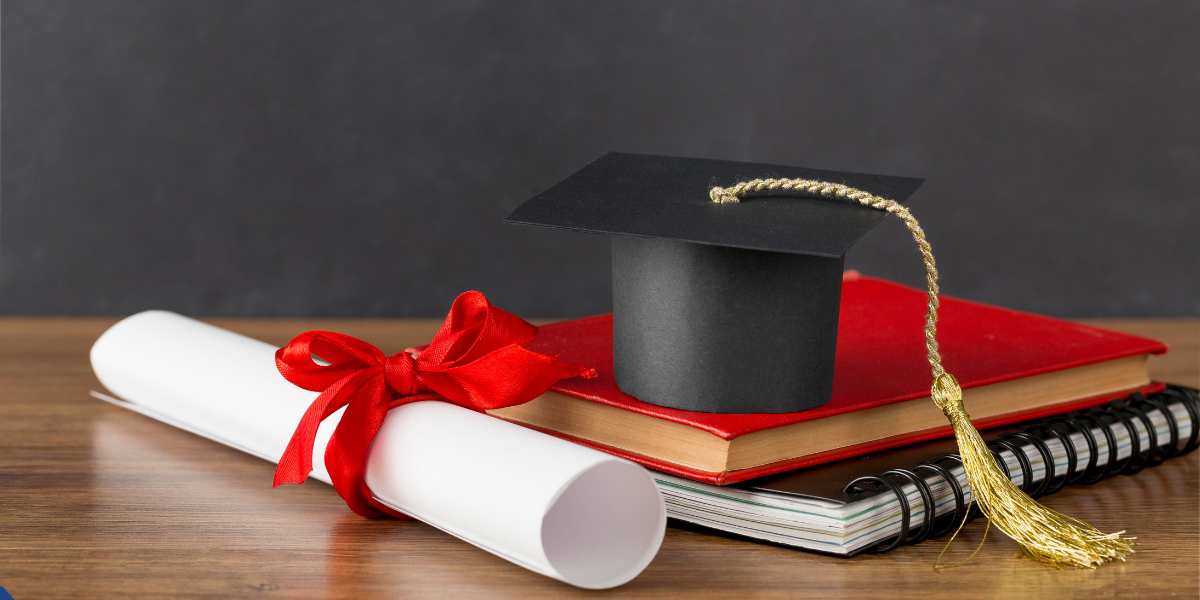 A black graduation cap with a golden tassel resting on top of a red book and spiral notebook, next to a rolled degree certificate tied with a red ribbon on a wooden table.