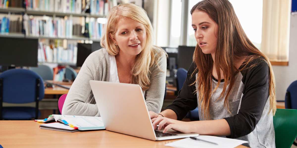 An educational consultant or teacher sitting with a young female student in a library, looking at a laptop screen together and discussing university options.