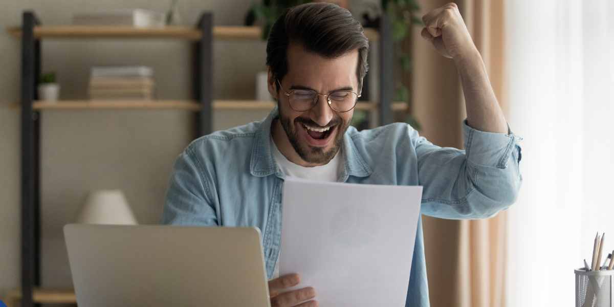 A happy male student cheering with excitement while looking at a university acceptance letter and a laptop, celebrating a successful UCAS Clearing application.