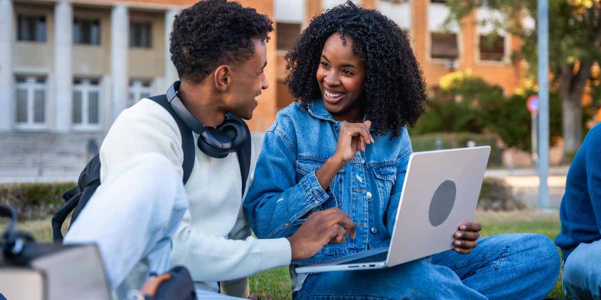 Two university students sitting on a campus lawn, smiling and looking at a laptop to research open course places during the UCAS Clearing process.