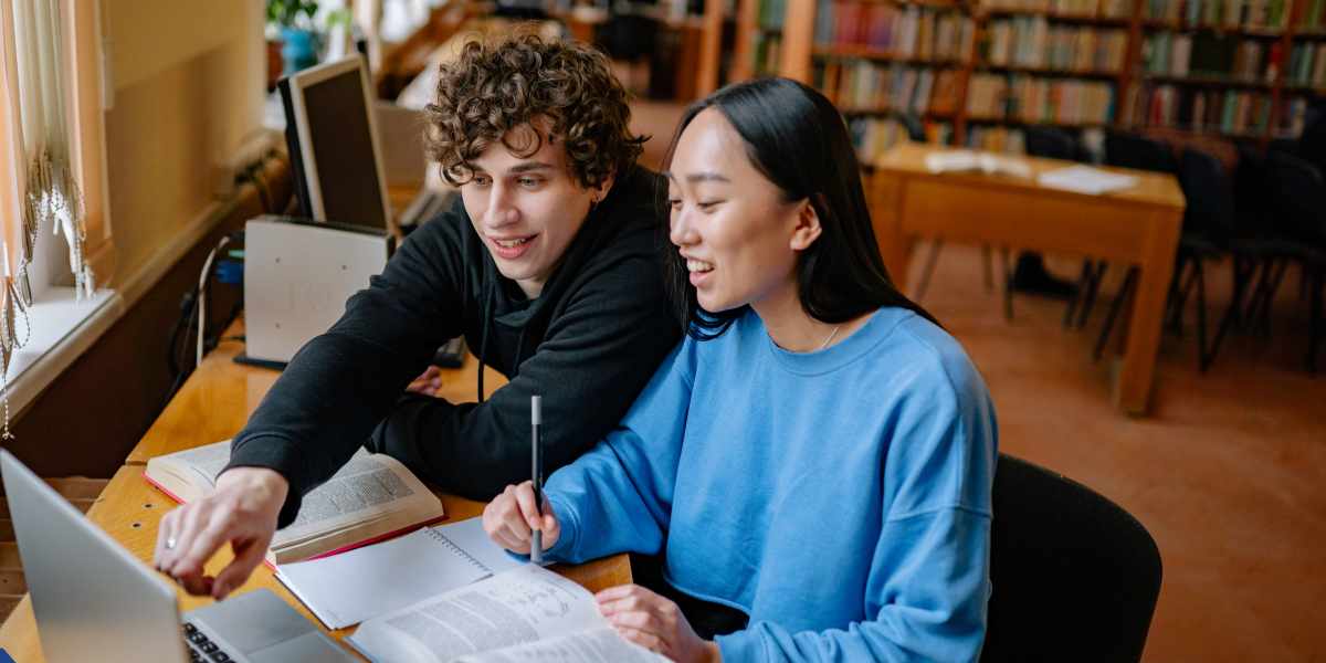 A diverse group of four young students—two male and two female—sitting closely around a wooden table in a well-lit library or classroom setting. They are leaning in together, smiling and cheerfully collaborating on their studies. Open binders, notebooks, pens, and a silver laptop are spread across the table in front of them, with bookshelves blurred in the background.