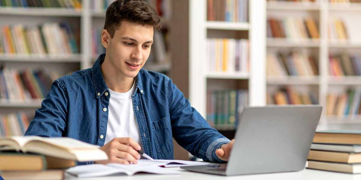 A young male student sitting at a white desk inside a bright, spacious library. He is smiling gently while looking at his laptop screen. In front of him is an open notebook where he is holding a pen, ready to take notes. Stacks of textbooks are placed on the desk beside him, with rows of bookshelves softly blurred in the background.