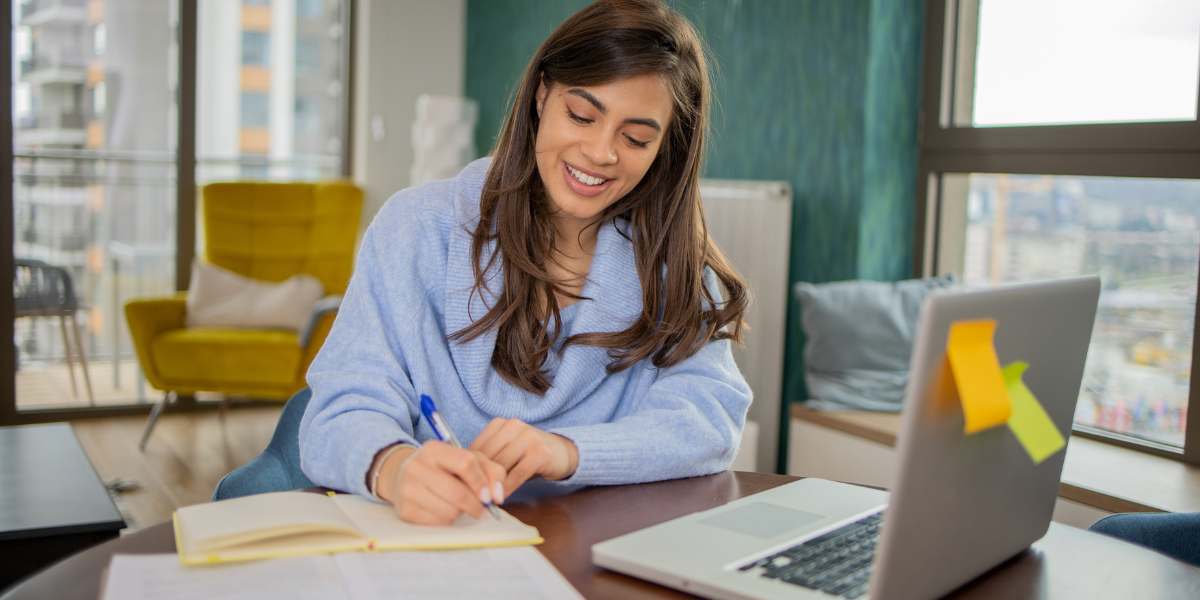 A smiling female student taking notes and using a laptop at home, representing online and distance learning degree options in the UK.