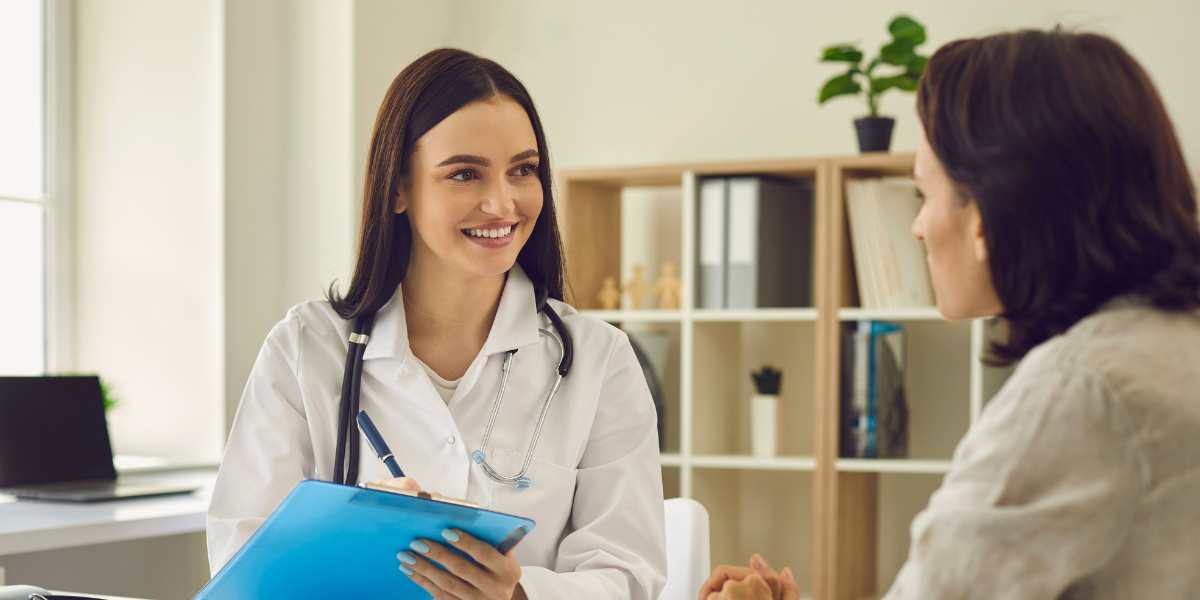 A smiling female doctor in a white coat with a stethoscope listening to a patient and holding a clipboard during a healthcare consultation in a bright office.