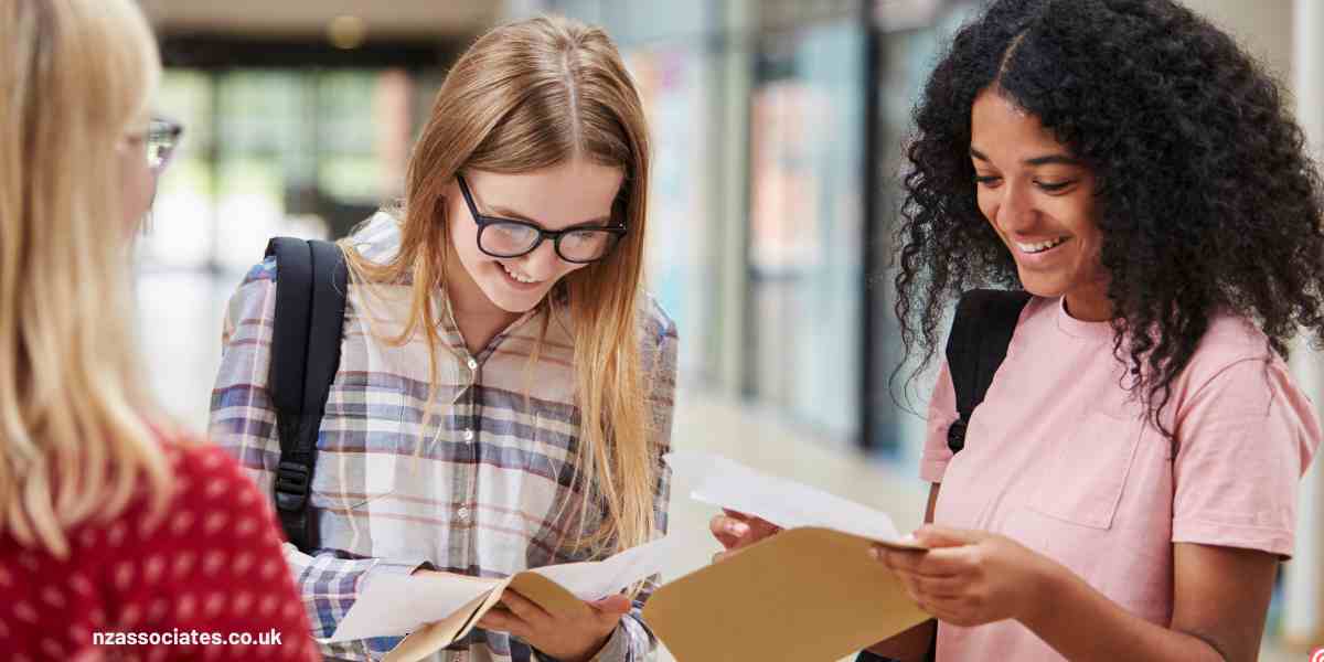 Two happy female students smiling as they open and check their A-Level exam results inside a UK college hallway.