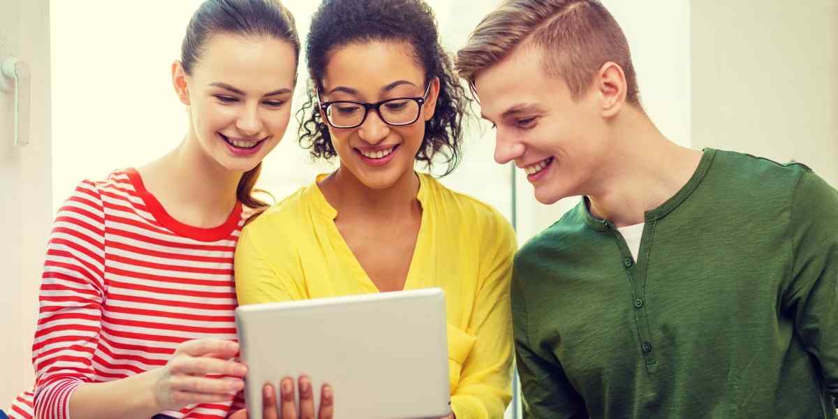 Three young diverse students smiling and looking together at a tablet screen, following the complete guide on how to apply for Student Finance England 2026/27.