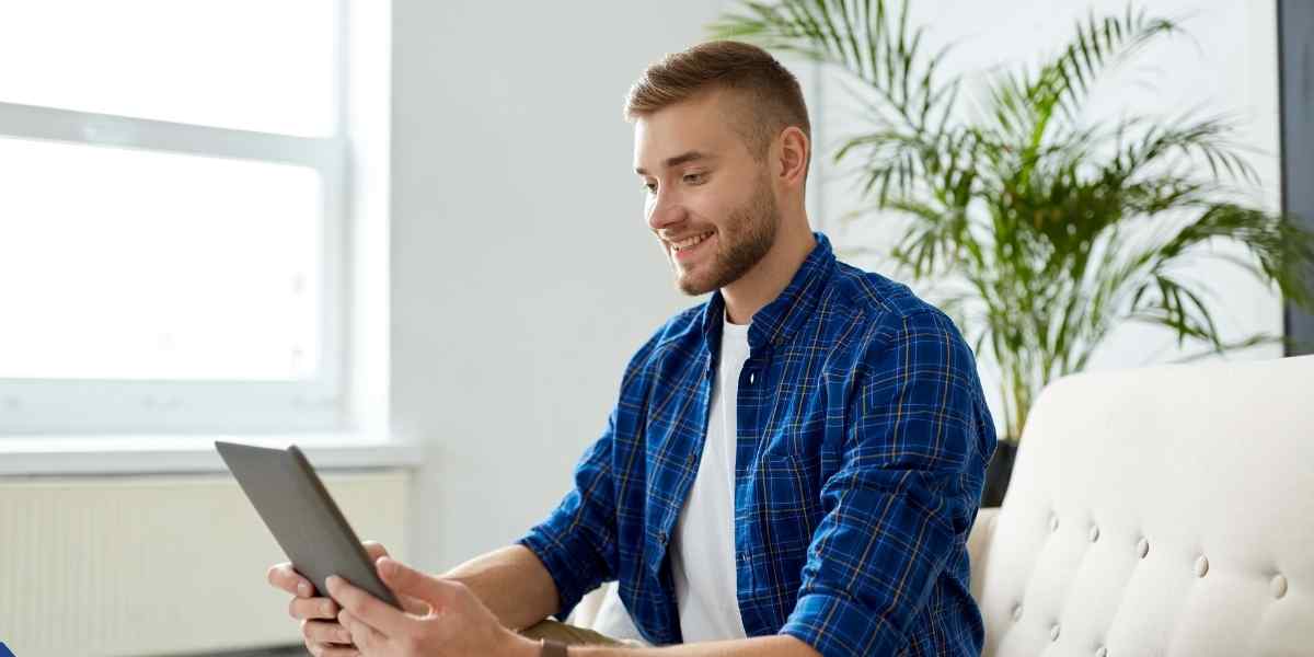 A young male student smiling while looking at his digital tablet on a sofa, researching how to apply for Student Finance England 2026/27 guide online.