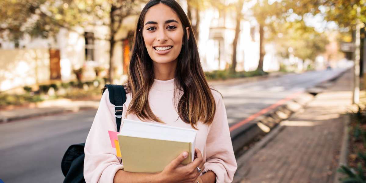 A young female student smiling confidently while holding her textbooks and wearing a backpack, standing on a sunny tree-lined street, representing a UK learner exploring their eligibility for the Lifelong Learning Entitlement (LLE)