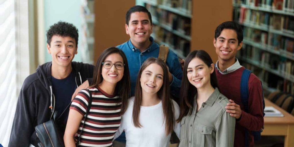 A diverse group of five smiling young adult students standing close together inside a library, looking directly at the camera with bookshelves visible in the background.