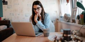 A young woman with glasses looking anxiously at her laptop screen at home, appearing worried about a missed deadline.