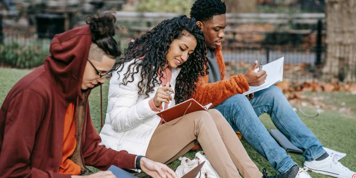 A group of diverse students sitting outside on the grass, reviewing their notes and planning their university finance applications.