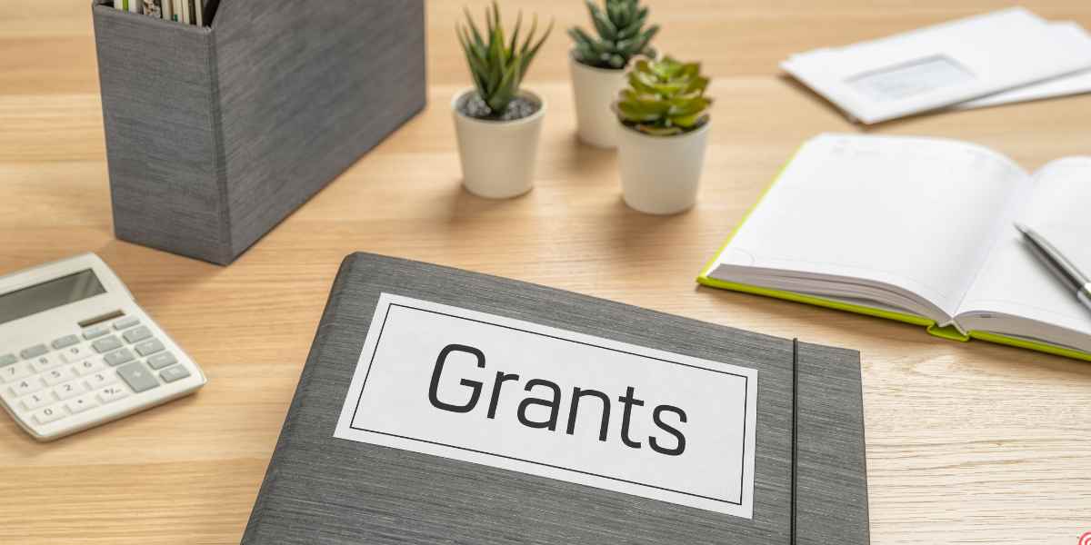 A close-up view of a folder labeled "Grants" alongside a calculator and open notebook on a wooden desk, illustrating extra financial support and institutional bursaries available for estranged students in the UK.