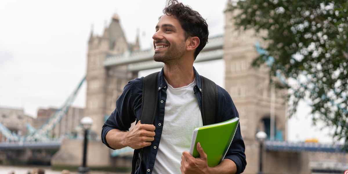A smiling young male student with a backpack holding a green folder, standing in front of London’s Tower Bridge, representing an independent journey through UK student finance for estranged students.