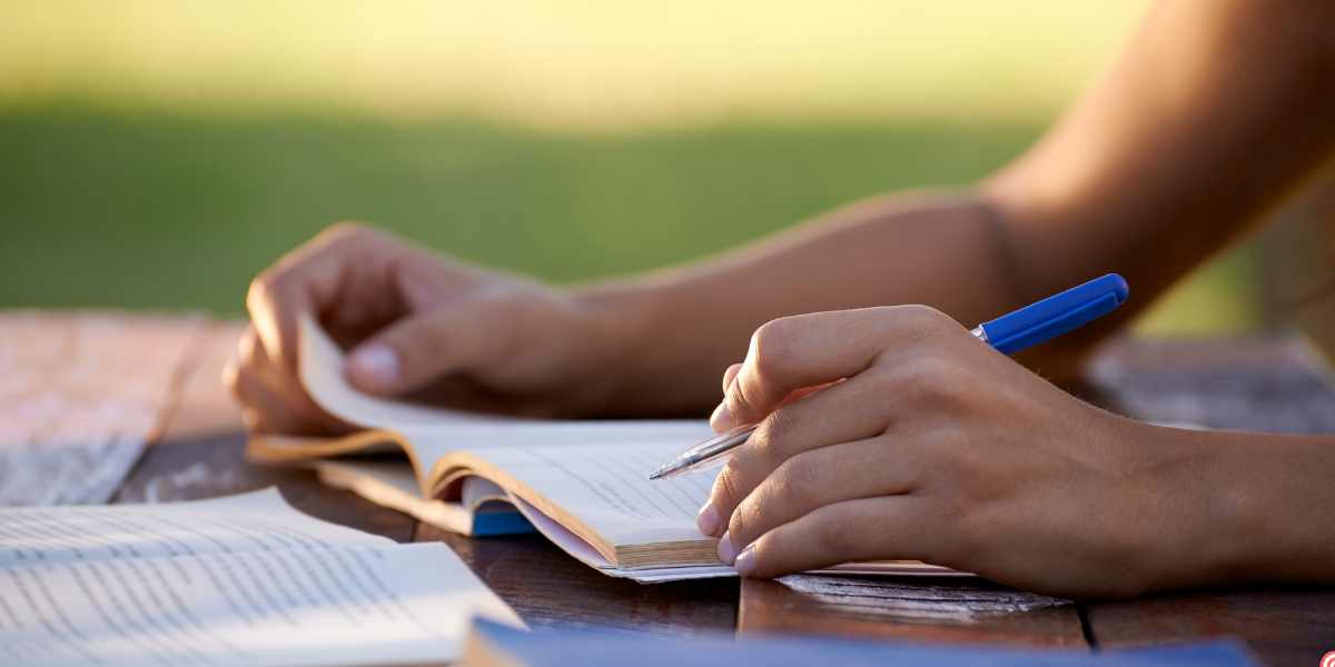 A close-up of a student's hands writing in a notebook and turning a page on an outdoor desk, representing academic preparation and study for alternative university entry routes like a UK Foundation Year.