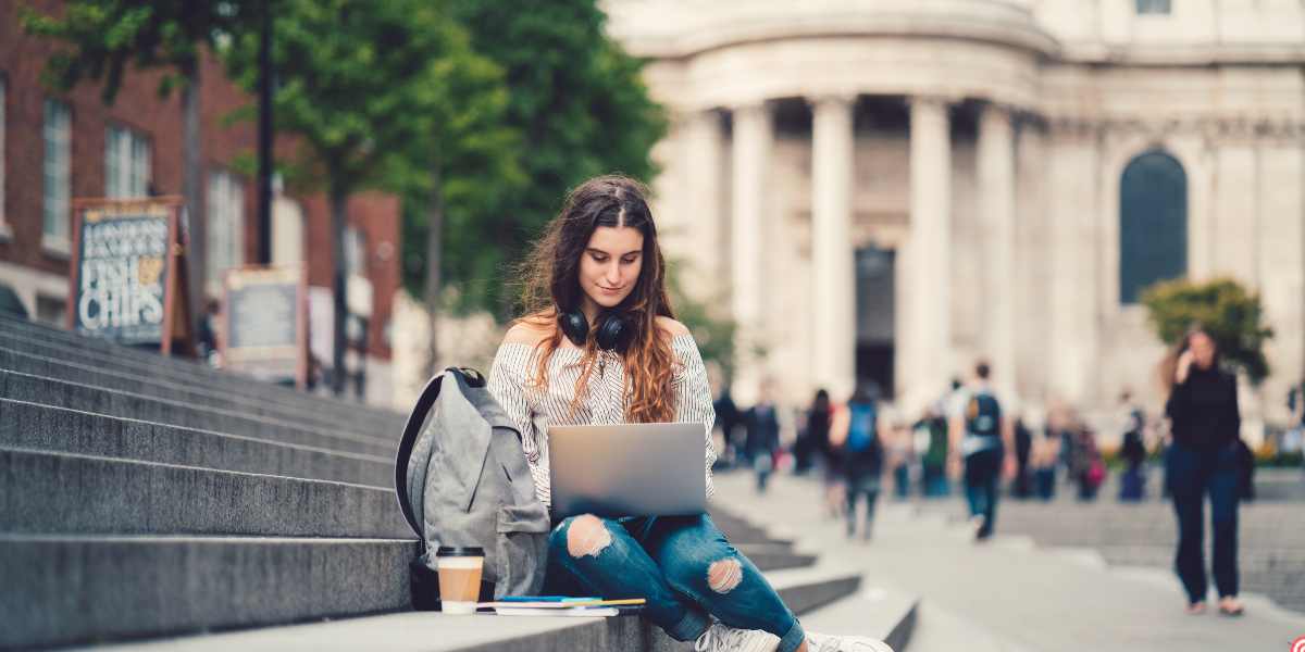 A female university student sitting on stone steps outdoors with her laptop, backpack, and a coffee cup, thoughtfully working on a bustling UK campus, illustrating whether investing in a UK university foundation year is worth it for long-term academic success.